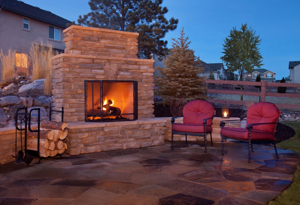 A backyard patio features a fireplace and casual furnishings. The outdoor fireplace burns with a bright orange glow and is covered by a metal grate. There is extra wood stored to the left of the stone fireplace in a black unit. There are two metal chairs with red cushions placed on the flagstone patio.